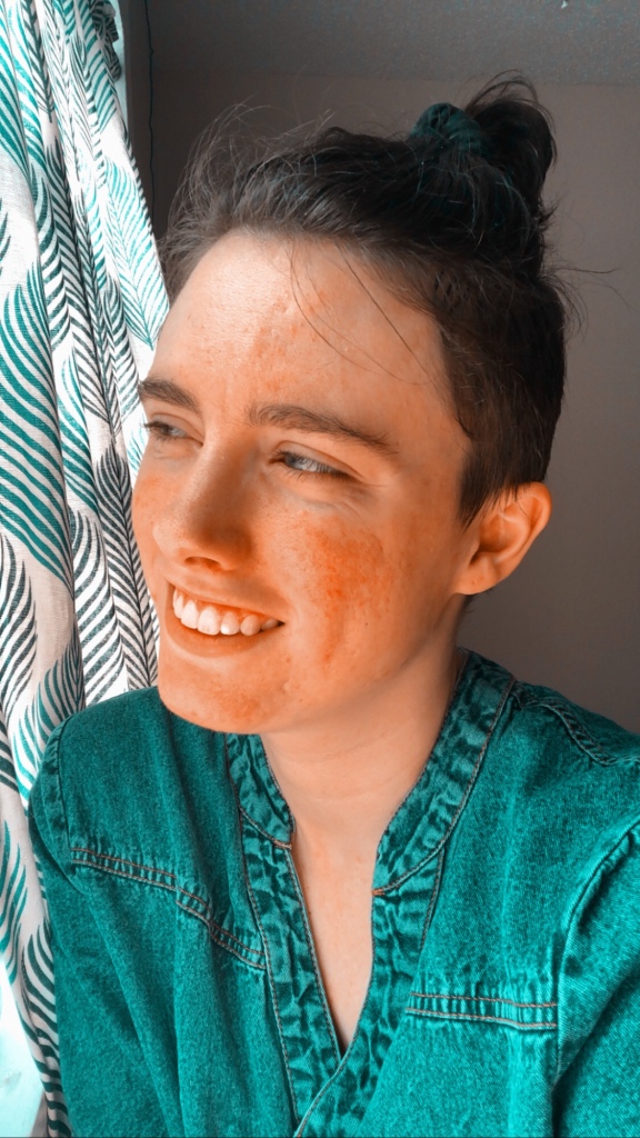 A young woman (me) sitting in front of white curtains with a pattern of dark green and grey leaves. She is smiling softly, wearing a v-neck denim dress with bronze buttons. Her hair is dark brown and the top part of her hair is in a bun, the sides are shaved. Baby hairs are everywhere. Her eyes are green.